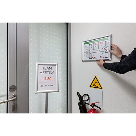 Eine Person befestigt einen Fluchtplan. Darunter ein Schild mit „Team Meeting 11.30“. Auf der Wand ein Warnzeichen und Feuerlöscher.