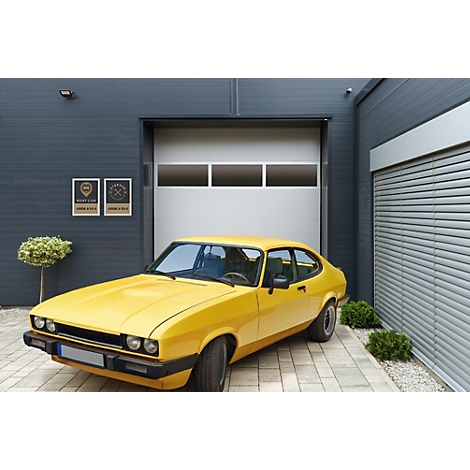 Ein gelbes Oldtimer-Coupé steht auf einer gepflasterten Einfahrt vor einem modernen Garagentor mit Lichtband.