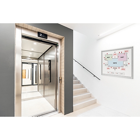 Moderne lift met open stalen deuren naast een trap en een evacuatieplan aan de witte muur.