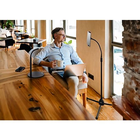 A man sits in an armchair with a laptop and a cup. A tablet is attached to a stand, the man is looking out of the window.
