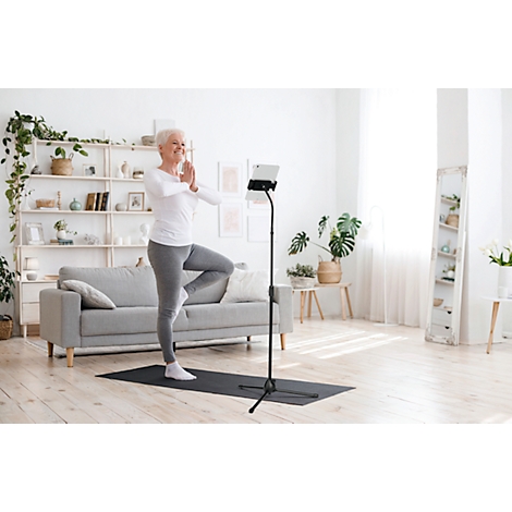 Senior woman doing yoga. She stands on one leg, arms in prayer position, in front of a tablet. Living room background.