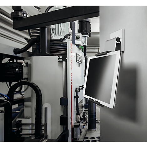 Tablet on holder in front of industrial plant. Bright display, black holder, gray wall.