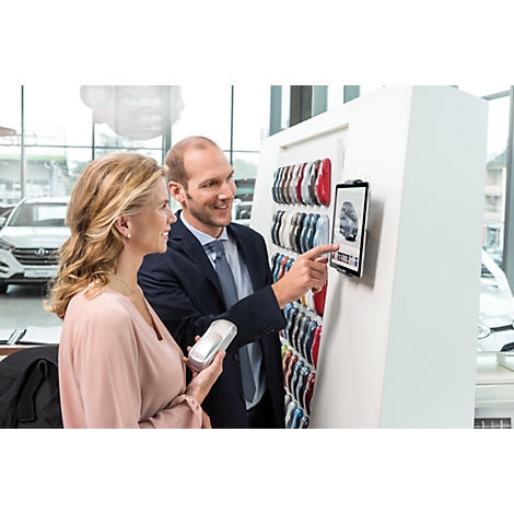 A car salesman shows a customer the color options for a car on a tablet PC. Cars are in the background.