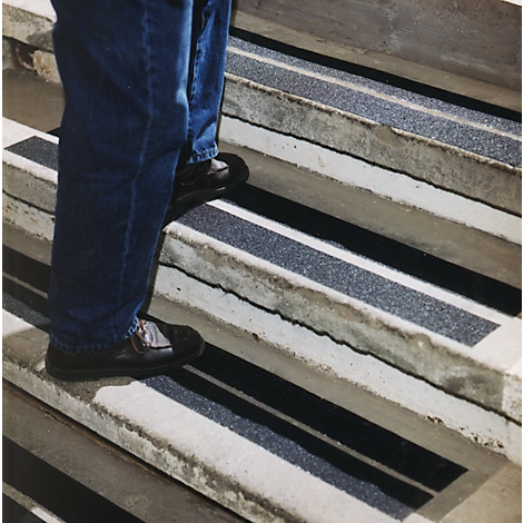 Person on stairs with non-slip strips. Black strips alternate with concrete. Jeans and black shoes.