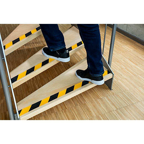 Person climbing stairs. Yellow and black warning stripes on the steps, light wood look, jeans and black shoes.