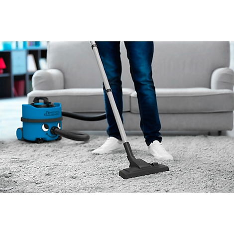 A person vacuums a carpet with a blue vacuum cleaner. A gray sofa is visible in the background.