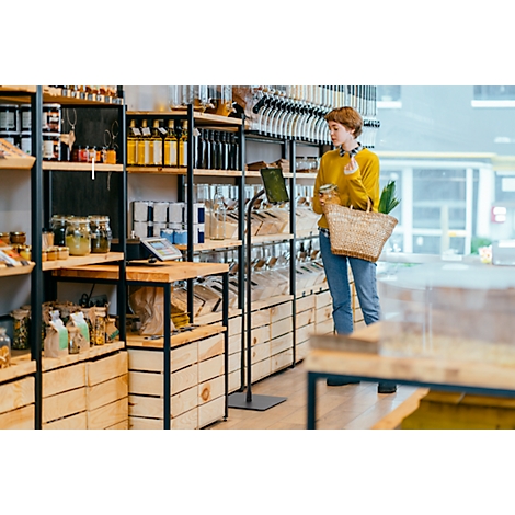 Jonge vrouw in een biologische winkel. Ze draagt een gele trui, jeans en een mandje. Schappen met voedsel.