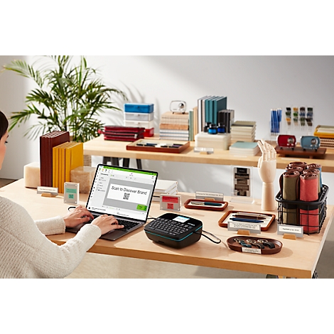 Frau am Laptop mit Etikettendrucker auf Tisch. Auf dem Bildschirm: "Scan to Discover Brand" mit QR-Code. Umgeben von Dekoartikeln.
