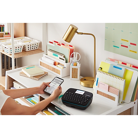 Een persoon die een labelmaker op een bureau gebruikt. Het bureau bevat notitieboekjes, pennen, ordners en een lamp.