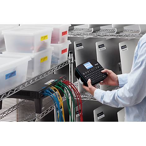 Persoon labelt computermonitoren met een labelmaker. Er zijn ook planken, bakken en netwerkkabels te zien.