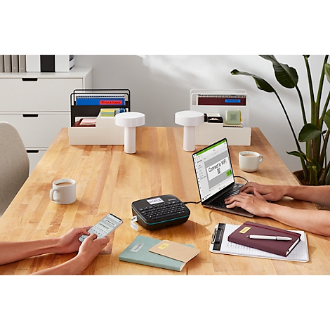 Kantoorscène: persoon achter laptop, anderen aan telefoon, labelmaker op tafel, koffie, pennen.