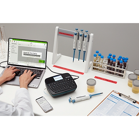 Une personne tape sur un ordinateur portable. À côté se trouvent des imprimantes d'étiquettes, des béchers, des tubes à essai et des pipettes.