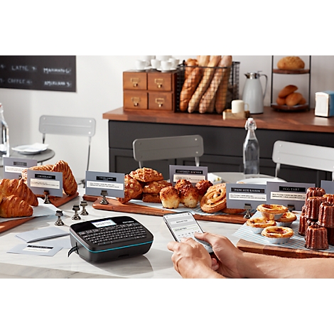 Une personne utilise un smartphone et une imprimante d'étiquettes sur une table avec des produits de boulangerie. Des panneaux affichent les noms des produits.