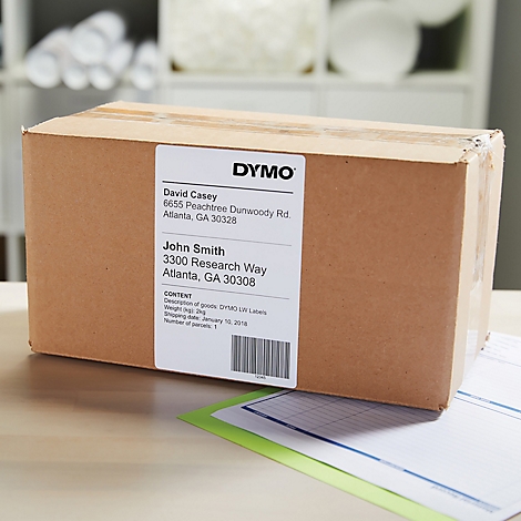 Boîte en carton marron avec étiquette DYMO, adresses et code-barres. D'autres documents sont sur la table.