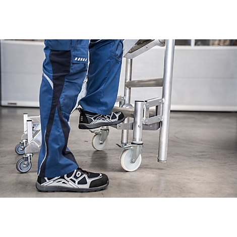 Personne debout sur une échelle à roulettes. Pantalon bleu, chaussures noires. Échelle en métal, fond gris.