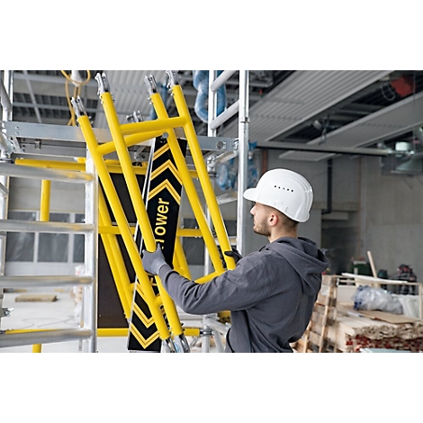 Un ouvrier avec un casque tient une partie d'échafaudage jaune. D'autres parties d'échafaudage et un chantier en arrière-plan.