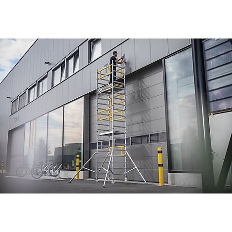 Un homme sur un échafaudage travaille sur un mur de bâtiment gris. Vélos au premier plan.