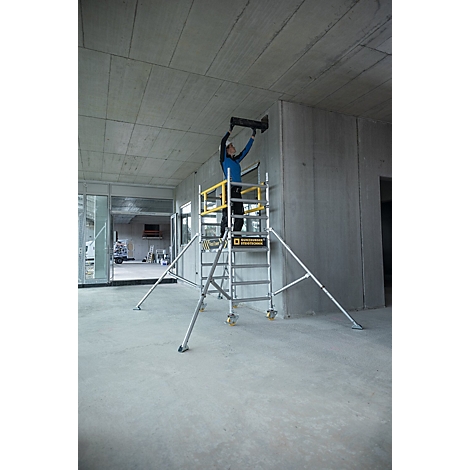 Une personne se tient sur un échafaudage et fixe un objet à un mur en béton. L'échafaudage a des éléments jaunes et se trouve dans un bâtiment en construction.