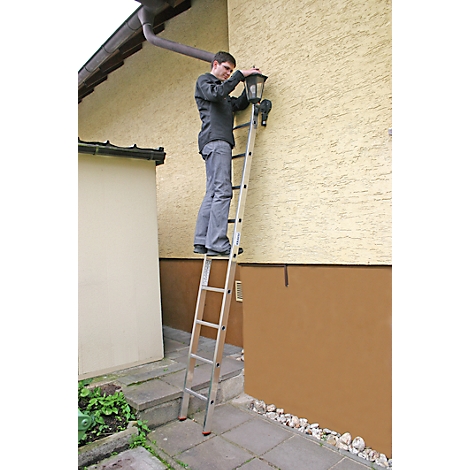 Un homme sur une échelle fixe une lampe au mur de la maison. Mur de la maison beige, marron. Gouttière, trottoir, arbustes et gravier.