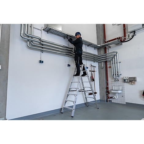 Un homme sur une échelle travaille sur des tuyaux sur un mur. En arrière-plan : plus de tuyaux, un évier et des outils.