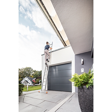 Un homme monte à une échelle sur un garage. Porte de garage grise, mur blanc, plantes vertes, ciel bleu.