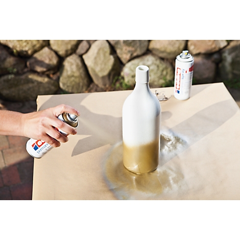 Een hand bespuit een witte fles met goudverf. Een andere spuitverfbus ligt op een oppervlak.