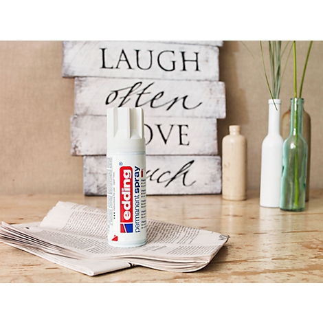 Een witte Edding permanente spuitbus staat op een krant, op de achtergrond houten borden met spreuken.