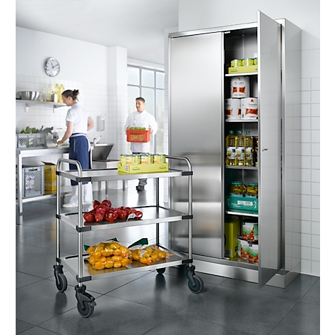 Küche: Edelstahl-Schrank, beladen. Zwei Köche arbeiten, Vorräte lagern. Tomaten, verpackt. Wandschrank offen.