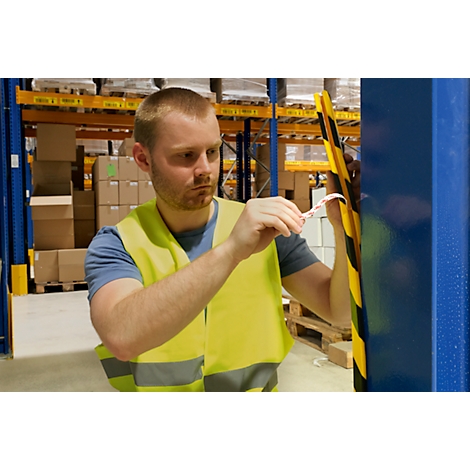 A man in a yellow safety vest attaches a tape to a warehouse pole. In the background, shelves with boxes are visible.