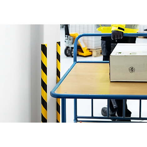 A worker pushes a cart with a cardboard box. Yellow and black warning poles, gray floor.