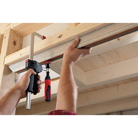 Een vakman klemt een werkstuk vast met een lijmklem. Lichtbruin hout, rode handgreep.