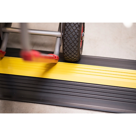 Close-up of a ramp with a yellow stripe and a wheelchair wheel. The ramp is black and the wheel has red accents.