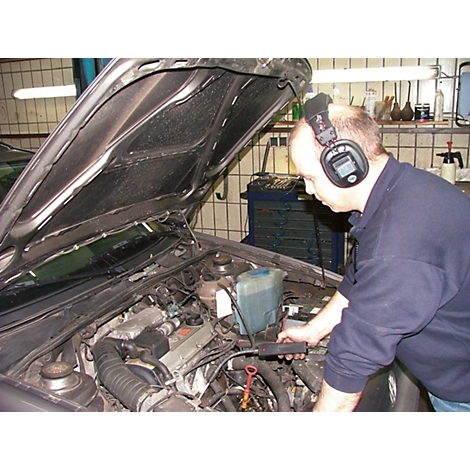 Ein Mechaniker mit Kopfhörern prüft einen Motorraum. Die Motorhaube ist geöffnet. Im Hintergrund Werkzeuge.