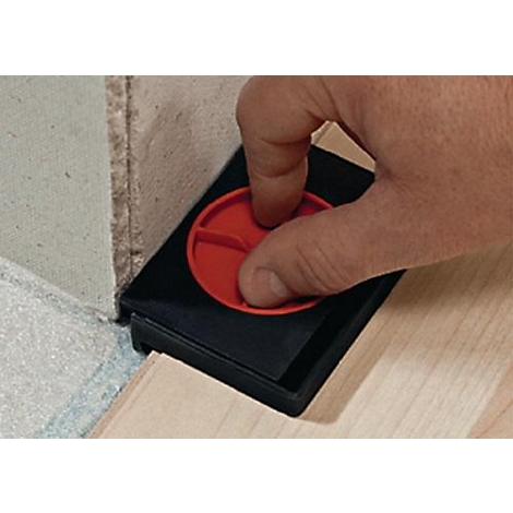 La main appuie sur un bouton rouge sur un outil noir, placé dans un coin. L'outil est entre le mur et le bois.