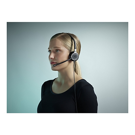Junge Frau mit blonden Haaren trÃ¤gt ein Headset mit Mikrofon. Sie blickt zur Seite, vor hellem Hintergrund.