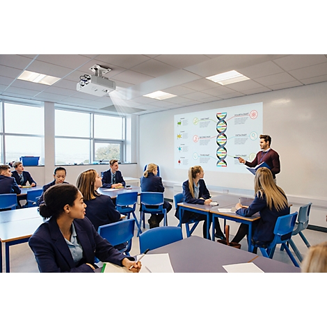 A teacher explains a DNA presentation in the classroom. Students sit at tables and listen. A projector projects the image.