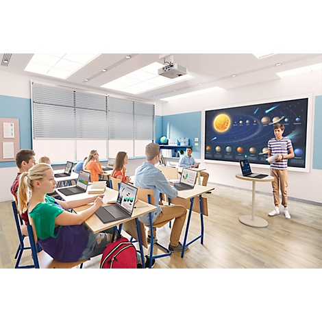 Students in classroom; a boy presents at the whiteboard; laptop on the table.