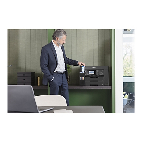 Ein Mann in Anzug befüllt einen Tintenstrahldrucker. Der Drucker steht auf einem Regal vor einer grünen Wand. Im Hintergrund ein Fenster.