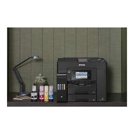 Ein schwarzer Epson-Multifunktionsdrucker steht auf einem Schreibtisch. Vor dem Gerät stehen Tintenflaschen. Links befindet sich eine Schreibtischlampe, rechts ein kleiner Blumentopf.