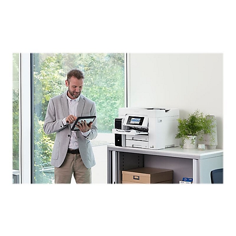 Un homme avec une tablette se tient devant une imprimante. L'imprimante est sur une armoire. Une fenÃªtre et des plantes sont visibles en arriÃ¨re-plan.