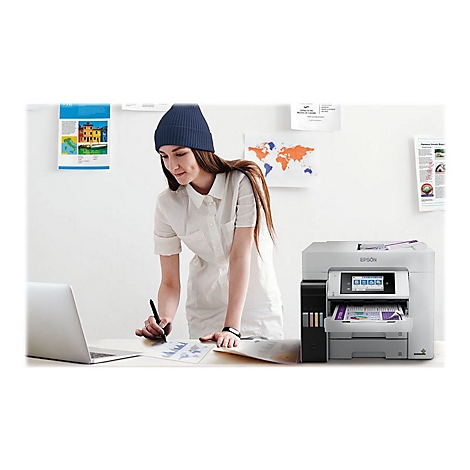 Une femme en chemise blanche et chapeau bleu se tient Ã  un bureau, avec une imprimante, un ordinateur portable et du papier, travaillant.