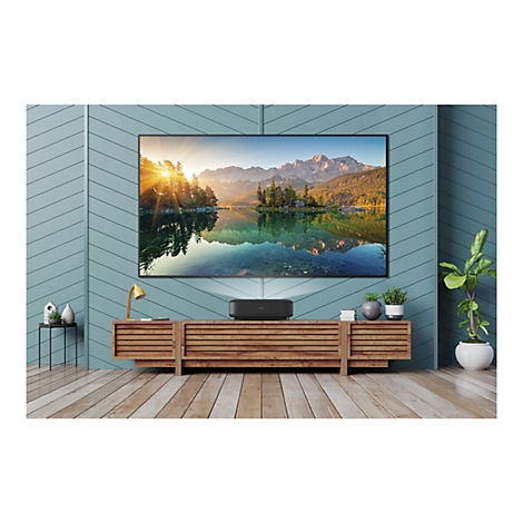 Großer Fernseher mit Seelandschaft. Darunter ein schwarzer Projektor auf einem Holzschrank. Zimmer mit Holzboden und grüner Wand.