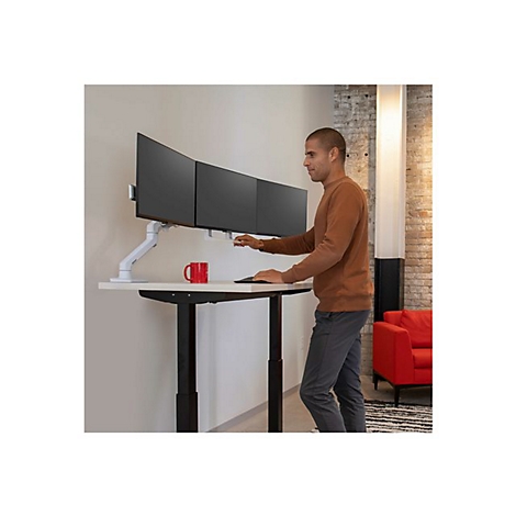Ein Mann steht an einem höhenverstellbaren Schreibtisch und bedient drei Monitore. Auf dem Tisch steht eine rote Tasse.