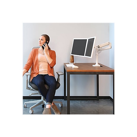 Eine Frau am Telefon sitzt an einem Schreibtisch mit zwei Bildschirmen. Sie trÃ¤gt einen orangefarbenen Cardigan, Jeans und Stiefel.