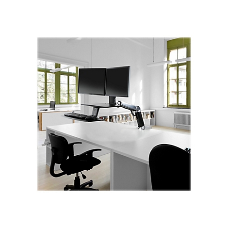 Büro mit zwei Monitoren auf einem verstellbaren Schreibtisch, Fenster mit grünen Rahmen.