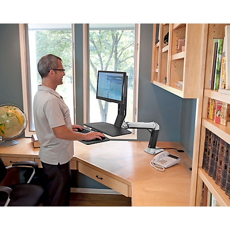A man stands at a height-adjustable desk and works on the computer, looking left. The office is bright and furnished.