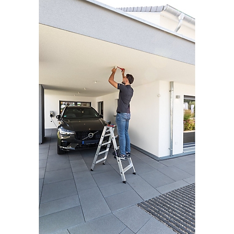Un homme sur une échelle installe un plafonnier au plafond d'un garage avec une voiture noire à l'intérieur.