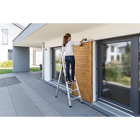 Une femme sur une échelle installe un écran d'intimité en bois. Le bâtiment a de grandes fenêtres et des carreaux gris.