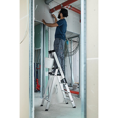 Homme sur une échelle lors de travaux de rénovation. Il peint un mur, avec des tuyaux et des câbles en arrière-plan.