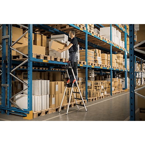 Un homme debout sur un escabeau soulève une boîte d'une étagère dans un entrepôt.